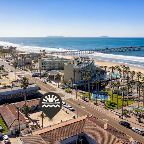 A coastal cityscape with a beach, pier, and buildings under a clear blue sky, featuring palm trees and a location pin icon.