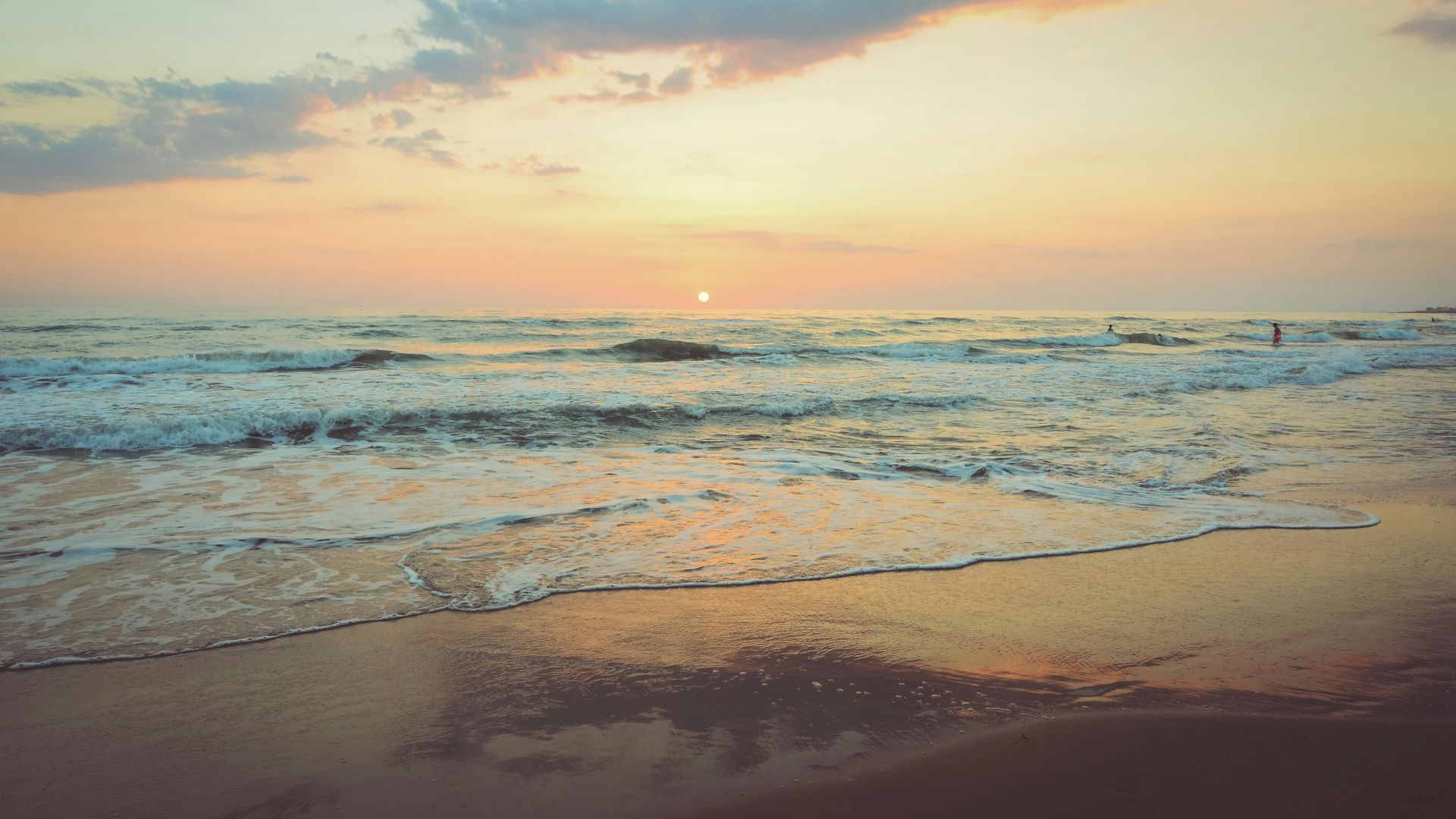 A serene beach at sunset with gentle waves lapping against the shore and a colorful sky with clouds illuminated by the setting sun.