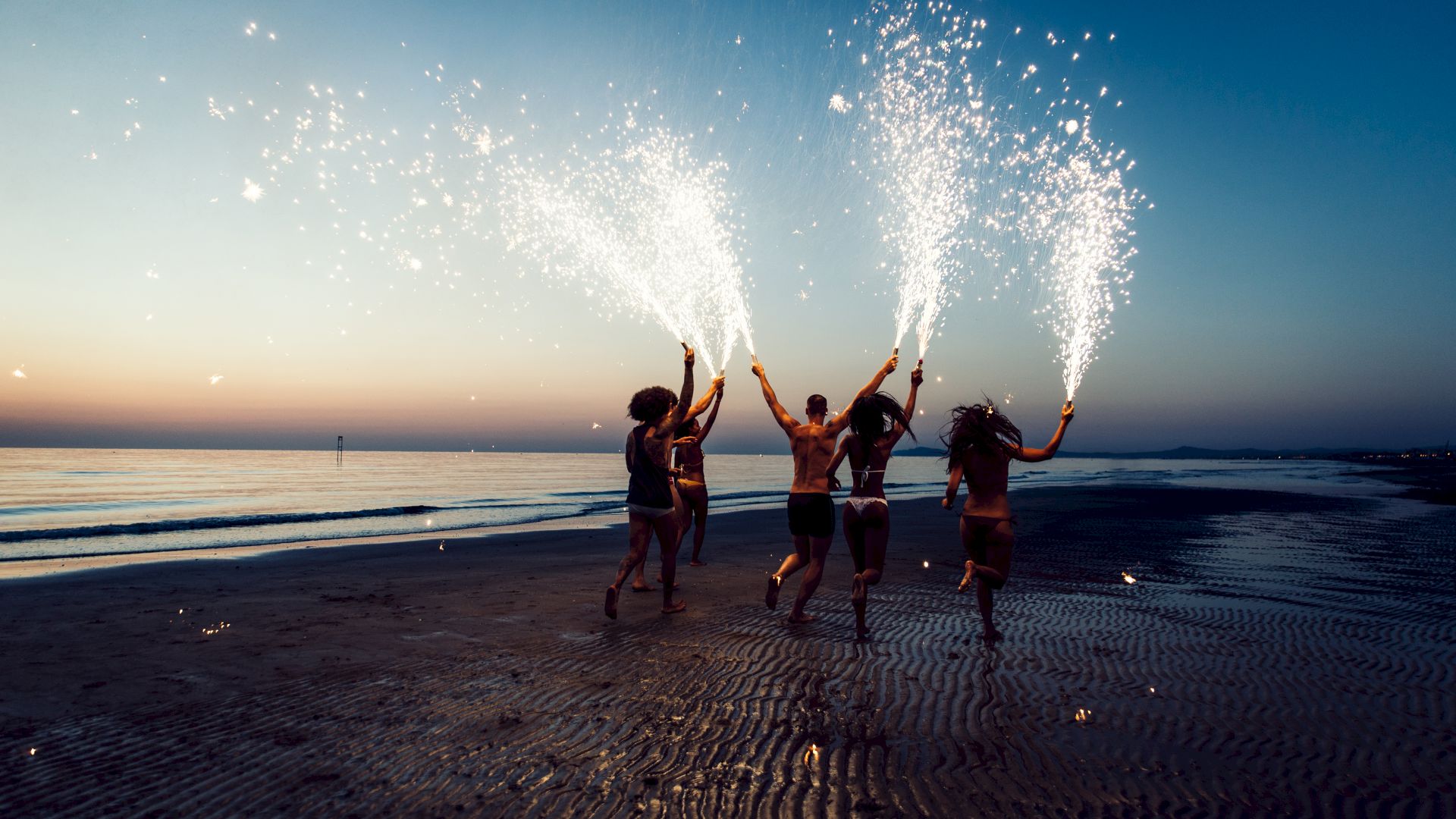 A group of people on a beach at sunset are holding sparklers, creating a festive and lively atmosphere on the sandy shore.