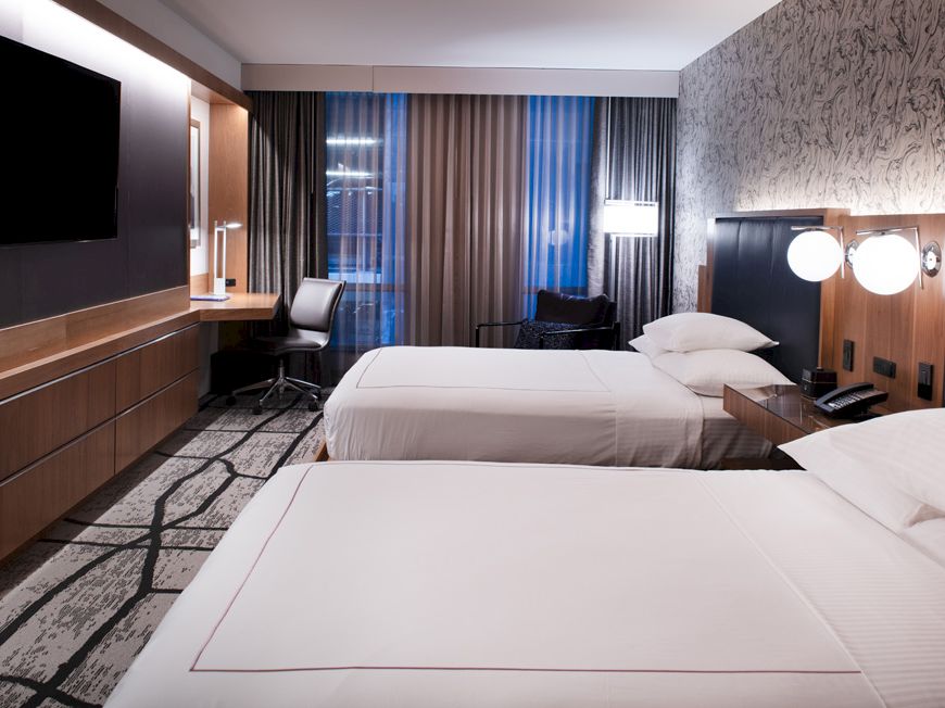 A modern hotel room with two double beds, a wall-mounted TV, a desk, lamps, and large windows with curtains, featuring contemporary decor and lighting.