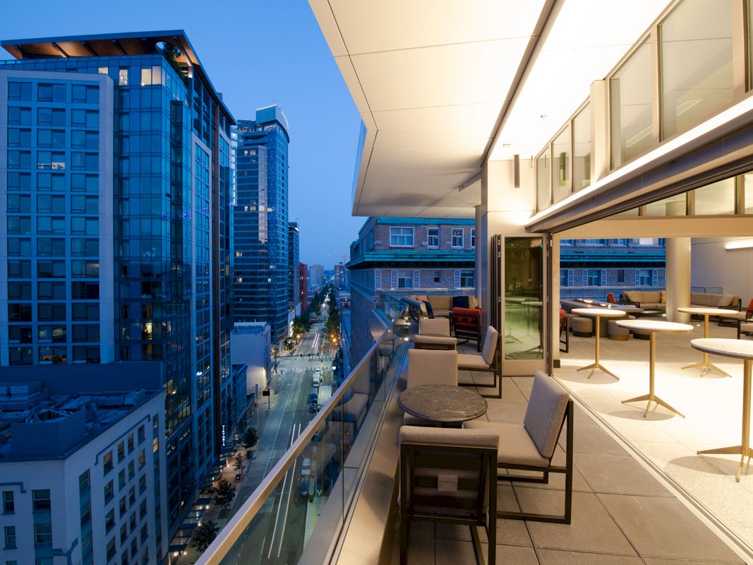 A modern balcony overlooking a city street with tall buildings, furnished with tables and chairs under a partially covered area.