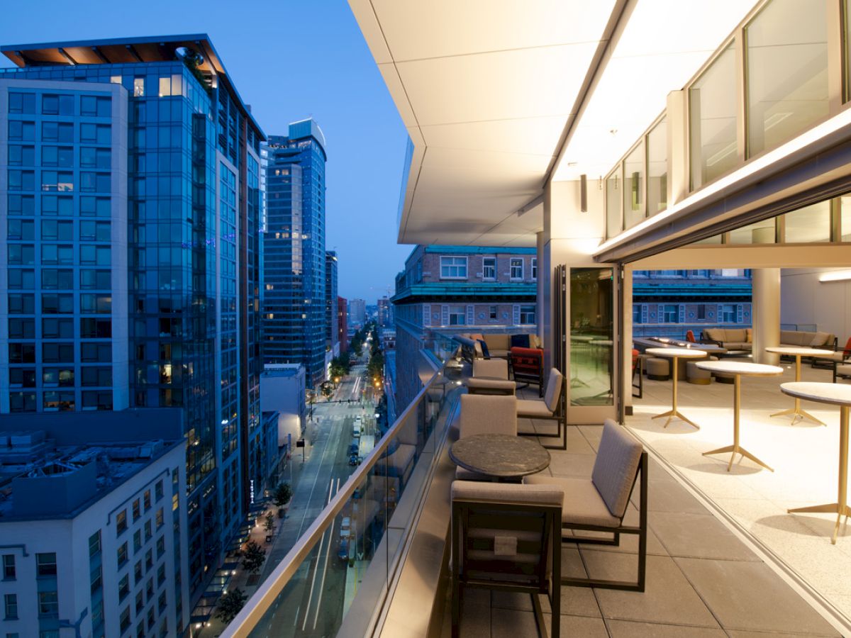 A modern cityscape at dusk featuring tall buildings and a balcony with tables and chairs overlooking a busy street below.
