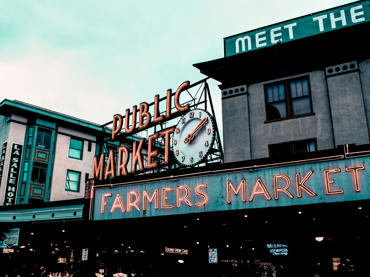 A neon sign reads "Public Market Center" with a clock and "Farmers Market" below. Buildings in the background, including one labeled "Parcel Hotel."