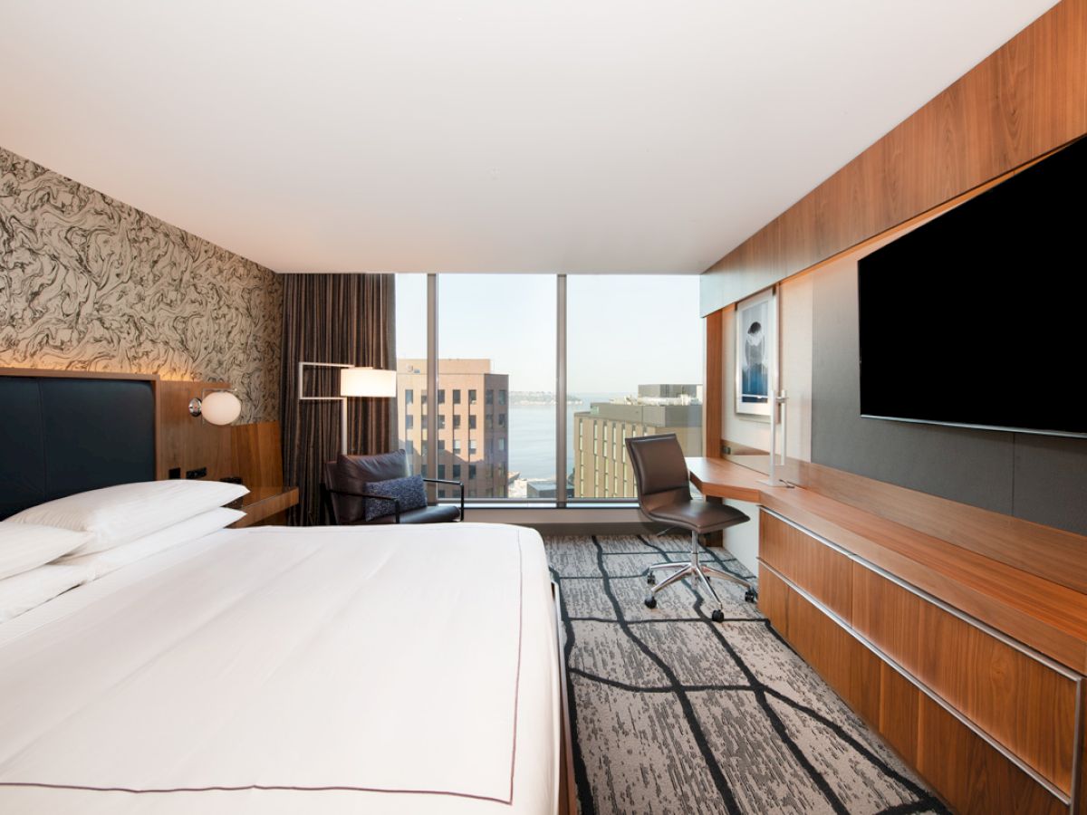 A modern hotel room with large windows, a bed, a desk with a chair, a TV, and a cityscape view in the background.