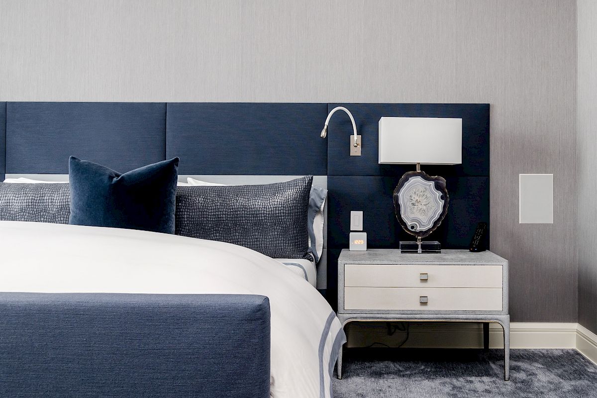 A modern bedroom with a blue and white theme, featuring a bed with blue headboard and pillows, a nightstand with a lamp, and a decorative clock.