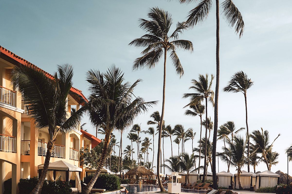 A serene poolside scene with lounge chairs and tall palm trees, adjacent to a resort building, under a clear sky.