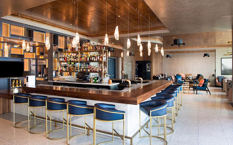 This image shows a modern bar with a wooden countertop, pendant lights, shelves of liquor bottles, blue bar stools, and lounge seating in the background.
