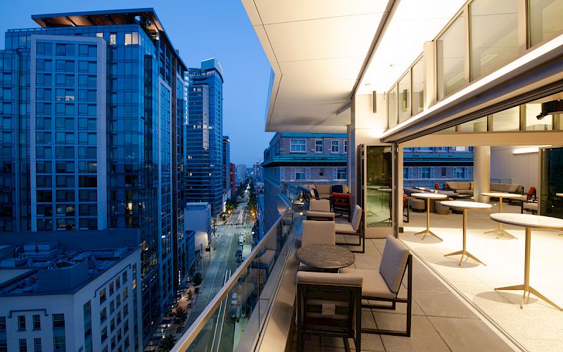 The image shows a modern balcony with seating, overlooking a city street with tall buildings during twilight.
