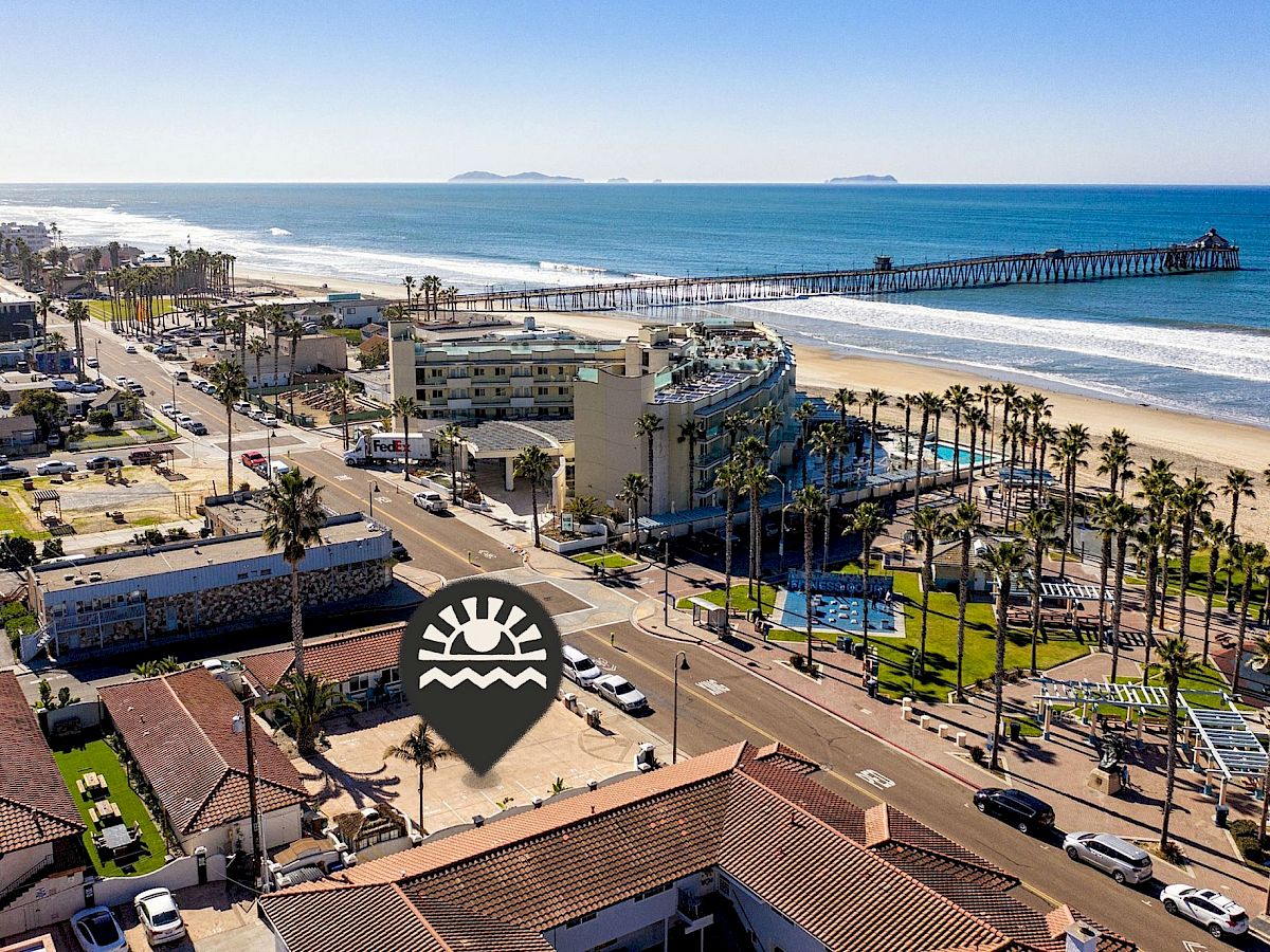 A coastal cityscape with a beach, pier, palm trees, and buildings along a road. An icon indicates a specific location.